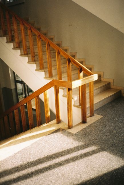 A woman carrying a large cardboard box up a narrow staircase during a home relocation process, with a man visible behind her. The stairs are carpeted and enclosed by white walls, with a black handrail