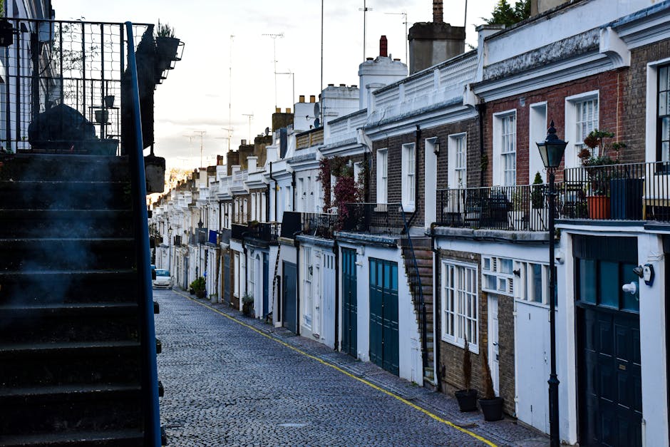 A row of typical London terraced houses with brick and stucco facades, each featuring small balconies with black metal railings, potted plants, and exterior lighting, situated along a narrow cobblesto