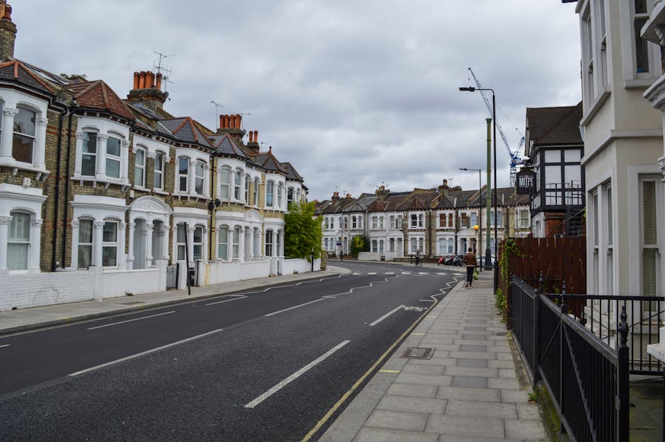 A row of typical London terraced houses with brick and stucco facades, each featuring small balconies with black metal railings, potted plants, and exterior lighting, situated along a narrow cobblesto